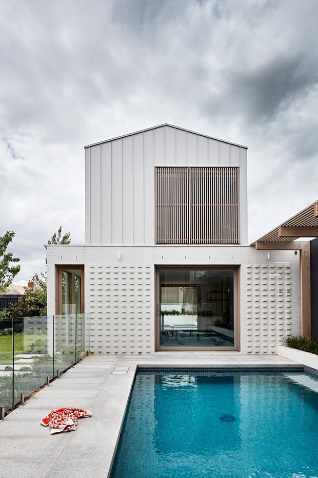 A contemporary two-storey residence viewed from the backyard, with a white vertically panelled upper volume, a textured breeze-block lower facade, timber screening over an upper opening, and large dark-framed glazing. A swimming pool and paved terrace occupy the foreground, with a glass pool fence and landscaped garden at the side.