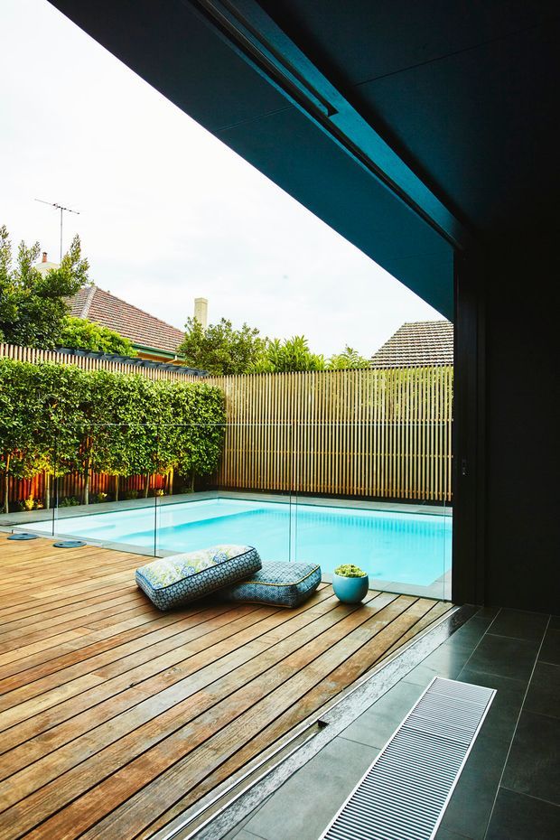 A contemporary covered outdoor entertaining area opens onto a small turquoise swimming pool. The space features a timber deck, frameless glass pool fencing, a vertical timber slat privacy fence, landscaped hedging, and patterned outdoor loungers. Dark ceiling and wall surfaces frame the view from the adjacent interior.