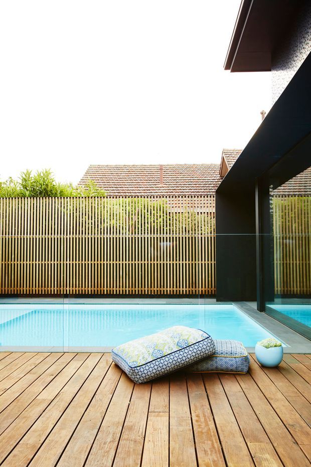 A bright outdoor pool area viewed from a timber deck. The turquoise swimming pool is enclosed by clear glass safety fencing and a vertical timber privacy screen, with a dark-framed glazed pavilion or building edge on the right. Patterned outdoor cushions and a small potted plant sit on the deck in the foreground. A tiled roof of a neighbouring or adjoining structure is visible beyond the screen.