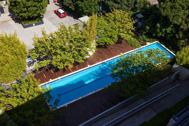 Aerial view of a long rectangular swimming pool surrounded by dark timber decking, mature landscaped trees and paved outdoor areas. The pool appears to be part of a private residential property, with surrounding fencing and access paths visible.