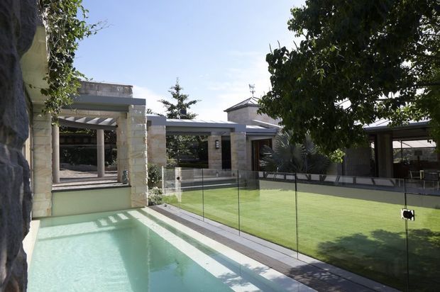 A modern landscaped residential courtyard viewed from beside a long narrow swimming pool. The pool is bordered by pale paving and frameless glass fencing, with a manicured lawn, mature trees, stone-clad pavilion structures, covered outdoor areas, and large glazed openings surrounding the space. Bright daylight and clean horizontal lines create a refined contemporary outdoor setting.