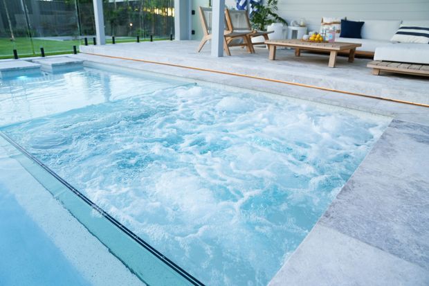 A bright indoor-outdoor pool area with a large bubbling spa or swim-jet section in the foreground, pale stone or concrete pool surrounds, and floor-to-ceiling glazing opening onto a lawn. A relaxed seating area with a timber coffee table, armchairs and a white sofa sits along the glazed wall, with light horizontal wall cladding behind it.