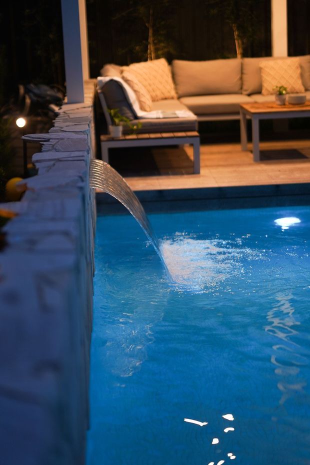 Evening view of a swimming pool with a curved wall-mounted water feature spilling into the blue water. A timber pool deck and an outdoor lounge area with cushioned seating and a low coffee table sit beyond the pool, framed by large dark windows and surrounded by a dimly lit wooded setting.