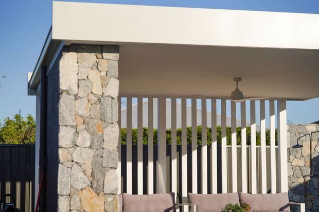 Exterior view of a contemporary covered outdoor seating area or patio, featuring a broad flat roof, pale rendered fascia, rough natural-stone columns and walls, vertical screening battens, and a ceiling-mounted pendant light. Neutral outdoor sofas are partially visible beneath the roof, with bright blue sky and landscaping beyond.