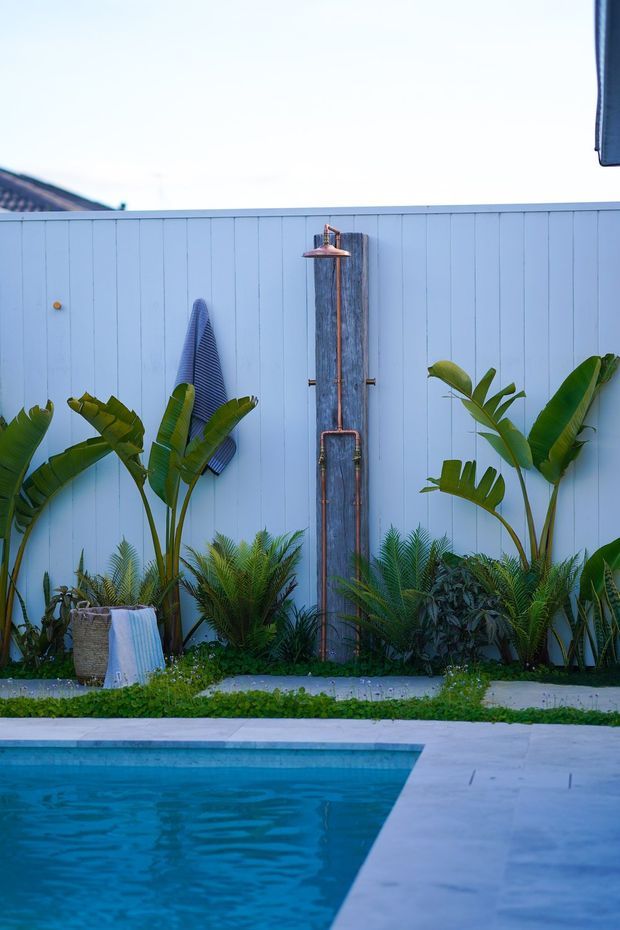 A tropical outdoor pool area with a blue-painted vertical timber privacy fence, lush palms and leafy planting, and a freestanding outdoor shower mounted on a weathered timber panel. A towel and decorative surfboard-like element add a relaxed resort feel, while pale paving surrounds the turquoise pool.