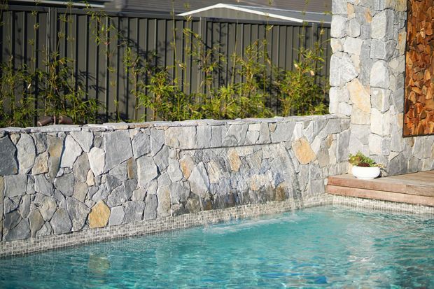 Close-up exterior view of a turquoise swimming pool bordered by a rough natural-stone retaining wall and raised stone pillar. A dark metal privacy fence with planted greenery sits behind the wall, while timber decking and a stacked wood feature are visible at the right edge.