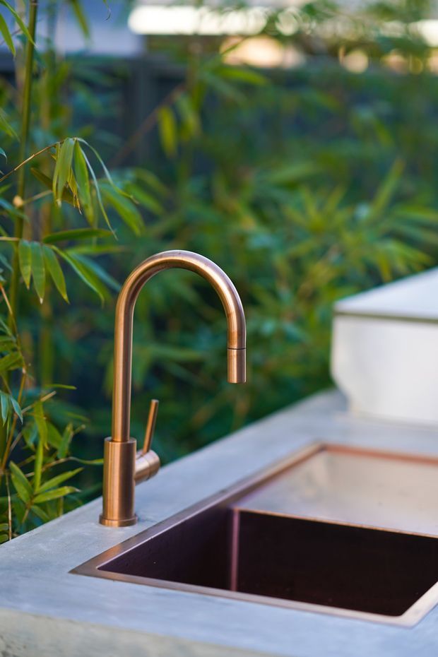 Close-up of an outdoor kitchen sink with a deep dark inset basin, pale stone benchtop, and curved copper-toned gooseneck tap. Dense green foliage forms the background, creating a natural outdoor setting.