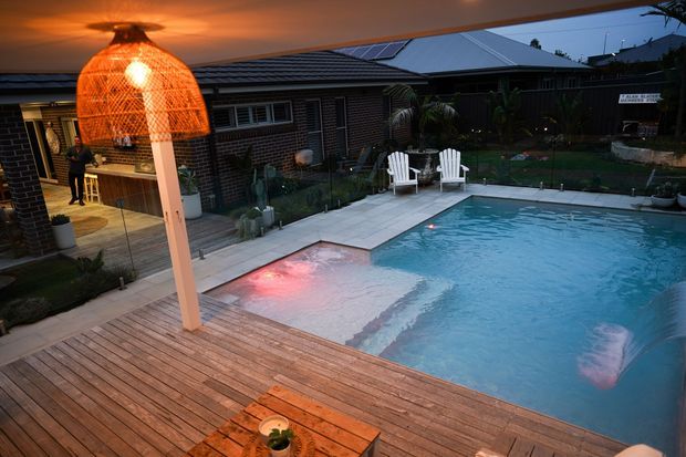 Evening view of a landscaped residential pool area with a large swimming pool, broad timber decking, pale paved surrounds, outdoor Adirondack-style chairs, and a prominent woven pendant light over the deck. A single-storey dark brick house with a low pitched metal roof and rooftop solar panels forms the backdrop.