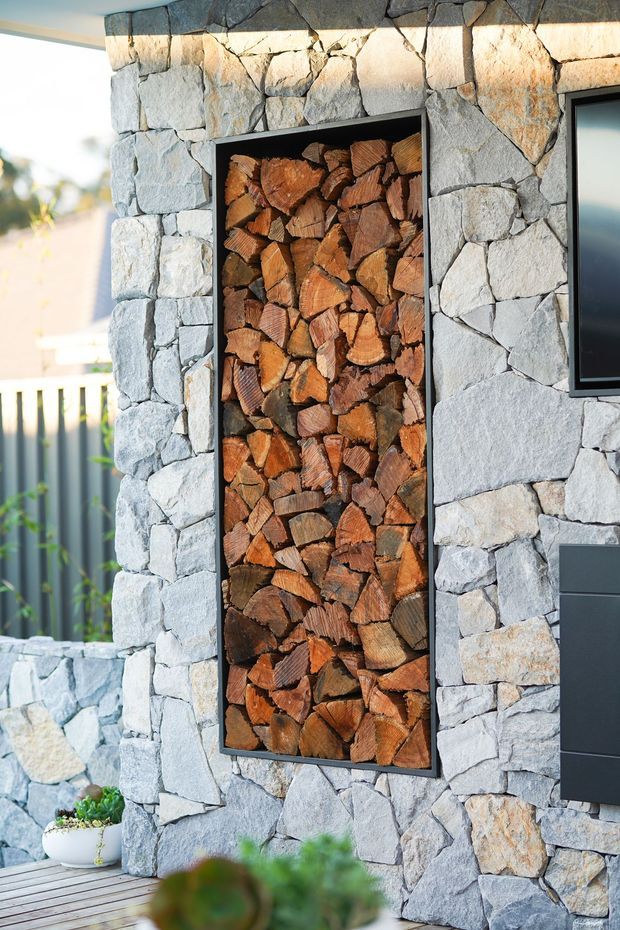 Close-up exterior view of a contemporary stone-clad wall featuring a tall recessed black-framed niche filled with neatly stacked firewood. A dark modern door and partial window are visible to the right, with timber decking and a small planter at the base. The rough, irregular grey stonework creates a strong natural feature facade.