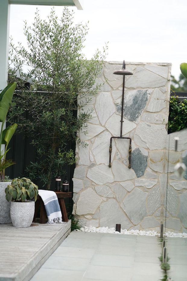 Outdoor shower area beside a pale irregular stone-clad privacy wall, with a dark metal shower fixture, pale tiled paving, timber decking, potted plants, hedging, and a striped towel. The scene has a natural, refined garden-courtyard feel.