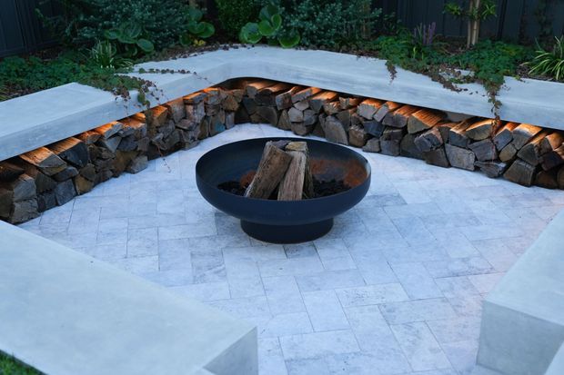 A landscaped outdoor courtyard or patio with pale stone paving, built-in angular concrete seating, and a circular black fire bowl at the centre. A low curved retaining wall faced with rough natural stone incorporates warm concealed lighting, with dense planting behind it.