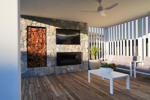 A bright, modern outdoor lounge area with a natural stone feature wall, wall-mounted television and fireplace, timber decking, white outdoor seating, a coffee table, vertical privacy screens, and a ceiling fan. The space has a relaxed indoor-outdoor character and strong afternoon sunlight.