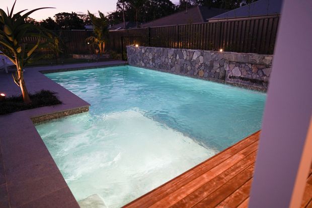 A landscaped residential pool area photographed at dusk, featuring a brightly illuminated swimming pool with a shallow ledge, warm timber decking, dark paved surrounds, tropical planting, a stone-clad retaining wall with subtle lighting, and a vertical timber privacy fence.