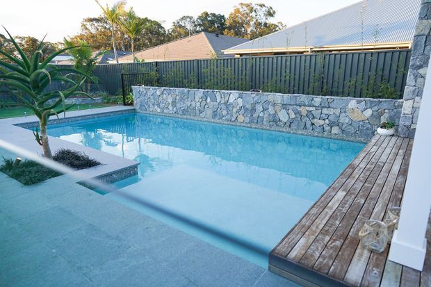 A contemporary backyard swimming pool with pale blue water, broad pale paving, and a timber-decked edge. A raised natural-stone retaining wall and dark vertical privacy fencing enclose the pool, with tropical planting and neighbouring rooflines visible beyond.