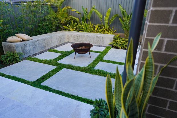 A compact landscaped courtyard with large pale concrete or stone stepping slabs set into grass, bordered by lush tropical planting and a low rendered bench wall with cushions. A shallow freestanding fire bowl sits centrally, while a dark grey vertical metal fence and brick building wall enclose the space.