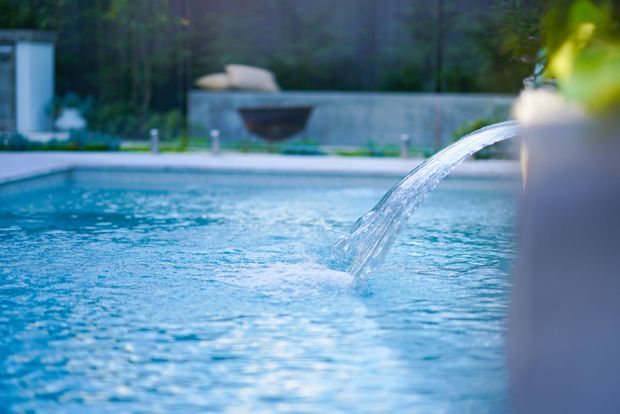 Close-up view of a blue swimming pool with a curved water spout flowing into the pool. A low concrete boundary wall, planted garden setting, and blurred outdoor features are visible in the background.