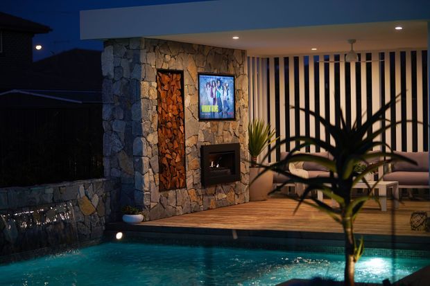 An evening view of a covered outdoor entertainment area beside a swimming pool. The space features a natural rough-stone feature wall with an inset fireplace and mounted television, timber decking, vertical privacy screening, lounge seating, recessed ceiling lighting, and tropical planting in the foreground. The illuminated pool forms the main foreground element.