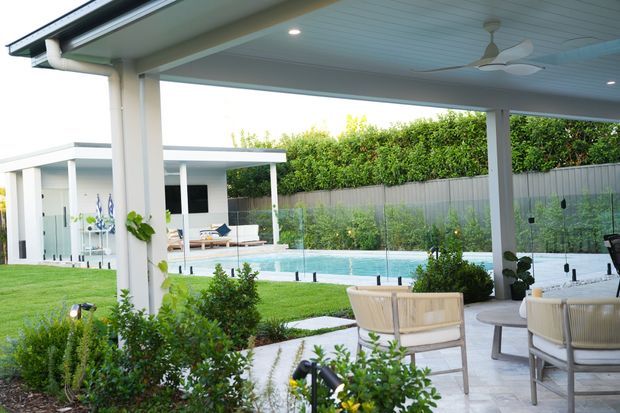 A bright covered outdoor entertaining area with a white-painted ceiling, recessed lighting, ceiling fan, and substantial supporting posts. It opens onto a landscaped yard and a rectangular swimming pool, with a paved patio furnished with light-coloured lounge chairs. A modern flat-roofed pavilion or pool house is visible beyond the lawn, screened by hedges and a tall boundary fence.