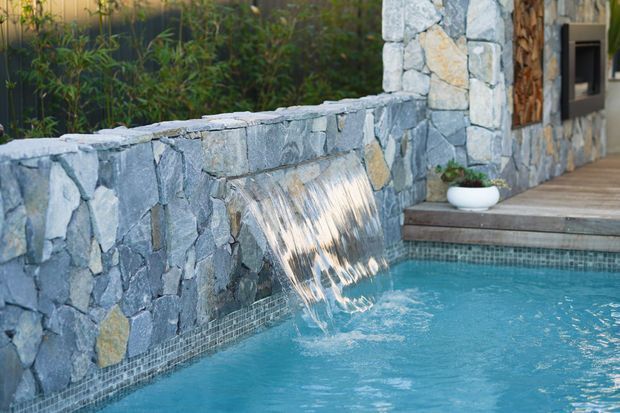 Close-up exterior view of a swimming pool beside a rustic stone-clad wall with an integrated rectangular water feature. Water spills into the turquoise pool, while timber decking, steps, planting, and part of a building entrance are visible in the background.