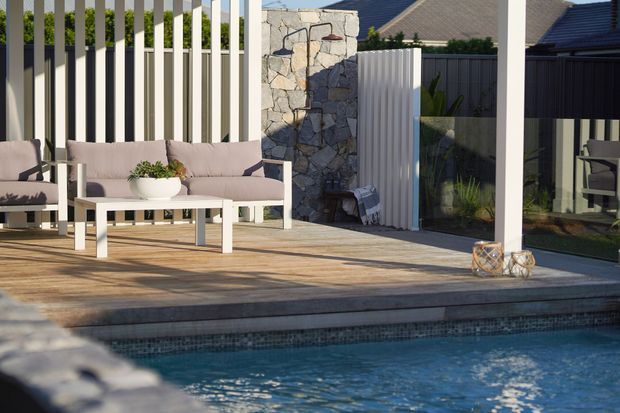 A contemporary outdoor poolside entertaining area with a timber deck, blue mosaic-tiled pool, pale outdoor lounge furniture, vertical privacy screening, a natural irregular-stone feature wall, and dark-framed glazing. The space is bright and relaxed, with layered textures and strong indoor-outdoor connection.