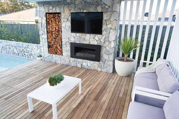 Outdoor entertaining area with a timber deck, pale cushioned seating, white coffee table, and a stone-clad feature wall incorporating a wall-mounted television, built-in fireplace, and recessed firewood storage. A swimming pool edge, privacy screening, and potted plant are also visible.