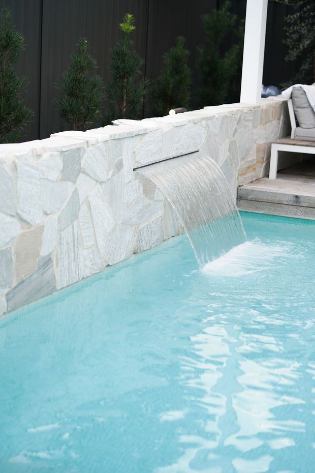 Close-up of a residential swimming pool with a curved sheet waterfall spilling from a pale irregular stone-clad wall. The pool water is turquoise, with dark vertical screening and evergreen planting behind the wall, plus a small outdoor seating area at the edge.