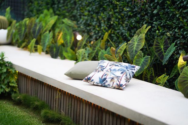 A landscaped outdoor seating area with a long built-in white bench or ledge set against dense tropical planting and a vertical green wall. The bench has a timber-clad vertical base and is styled with neutral and botanical-patterned cushions, creating a lush garden retreat.