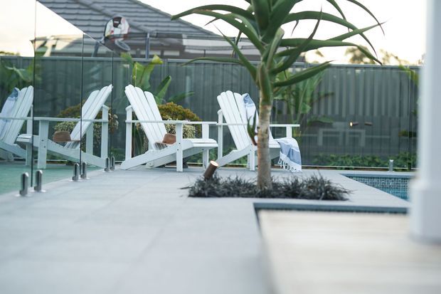 Contemporary outdoor pool area with a pale paved deck, several white Adirondack-style lounge chairs, a planted feature bed with a tall sculptural plant, and a glass pool fence. A residential building and boundary fencing are visible in the background.