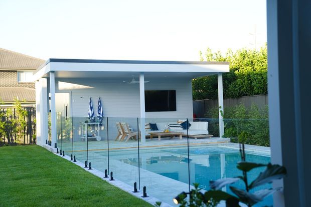 Contemporary poolside pavilion with a flat roof, white structural posts, glass pool fencing, and an outdoor lounge area furnished with seating and a wall-mounted television. The pavilion opens onto a bright blue swimming pool and landscaped backyard.