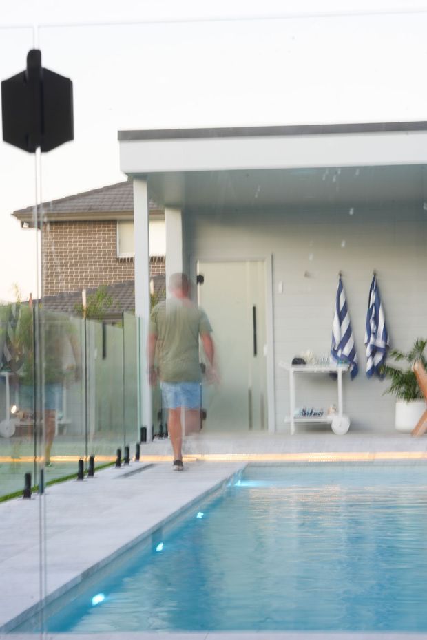 A contemporary outdoor swimming pool beside a modern pavilion or pool house. The pool is bordered by pale paving and frameless glass fencing with dark fittings. The pavilion has a flat roof, white rendered walls, a glazed or sheltered entry, built-in shelving, and striped pool towels. A neighbouring brick-tiled house is visible in the background, with a large black pendant light hanging in the foreground.