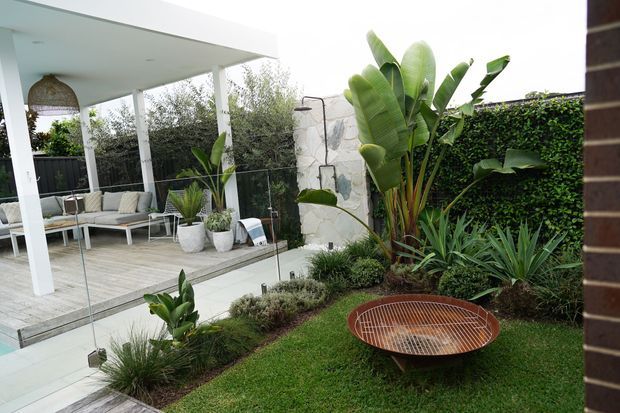 A landscaped residential courtyard viewed from beside a pool, featuring a covered outdoor lounge with white columns and a pale timber deck, a built stone feature wall with an outdoor shower, lush tropical planting, a clipped green privacy wall, and a circular rust-coloured metal fire bowl.