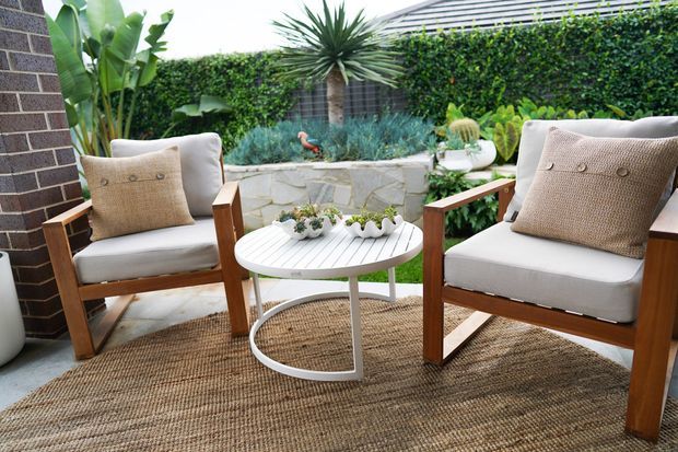 A styled outdoor seating area with two timber-framed lounge chairs and neutral cushions around a small round white table. A woven natural-fibre rug sits on a paved surface, with a low irregular-stone planter wall and dense tropical landscaping behind. A dark brick wall and tiled roof edge frame the courtyard-like setting.
