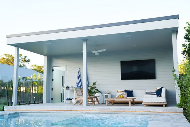 Bright modern poolside outdoor entertaining area with a flat-roofed covered pavilion, white horizontal wall cladding, slim structural posts, recessed ceiling lights and a ceiling fan. The space includes an outdoor lounge, coffee table, dining setting, wall-mounted television and direct views over a swimming pool.