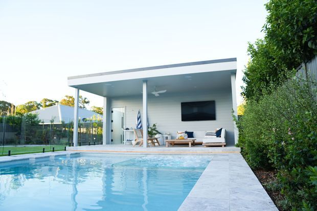 A modern poolside outdoor living pavilion with a flat roof, slim white structural posts, pale wall cladding, and an open entertaining area furnished with a sofa, chairs, coffee table, and wall-mounted television. A large rectangular swimming pool dominates the foreground, with light stone paving and landscaped screening around the perimeter.