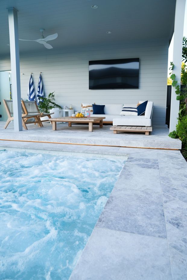A bright poolside outdoor living area with a covered pavilion, white horizontal wall panelling, ceiling fan, wall-mounted television, outdoor sectional sofa, timber coffee table, and woven lounge chairs. The foreground is dominated by a pale blue swimming pool with pale stone paving around it, while tropical planting frames the edges.