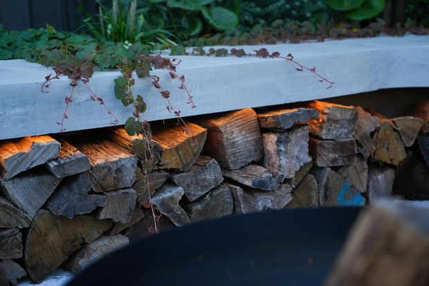 Close-up of a landscaped outdoor feature with a smooth concrete ledge above a stacked natural-stone retaining wall. Trailing plants spill over the edge, while warm lighting illuminates the wood-toned stonework and a dark curved surface is visible in the foreground.