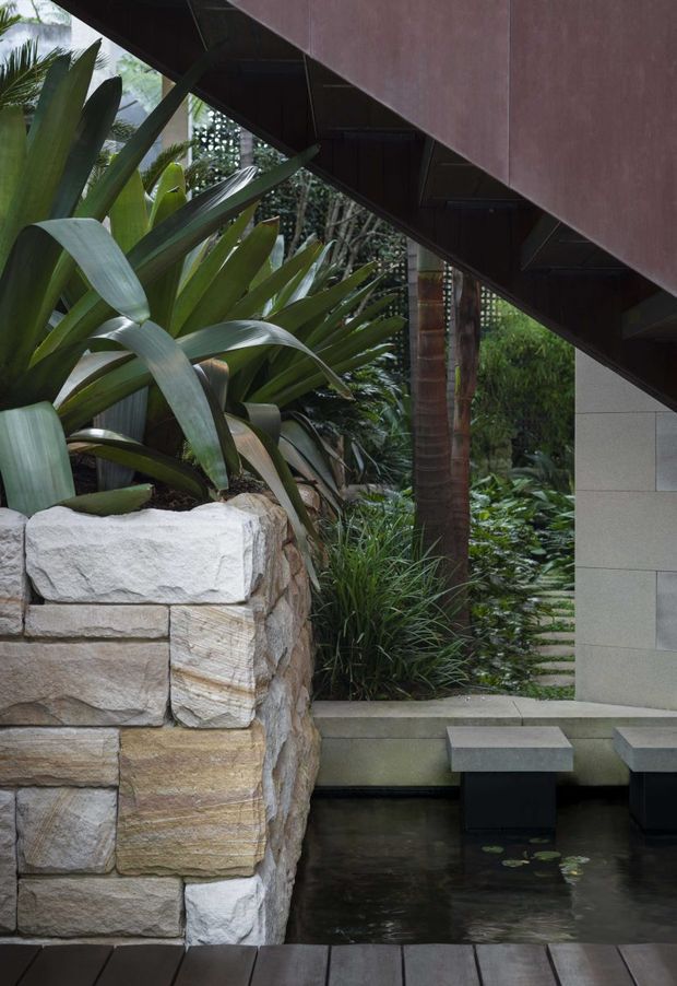 A lush landscaped courtyard or entry garden beside a contemporary building, featuring a shallow reflective pond with stepping stools, a large natural stacked-stone retaining wall, dense tropical planting, and a dark angular overhead roof or stair structure. The setting has a calm, secluded, resort-like atmosphere with strong integration of architecture and landscape.