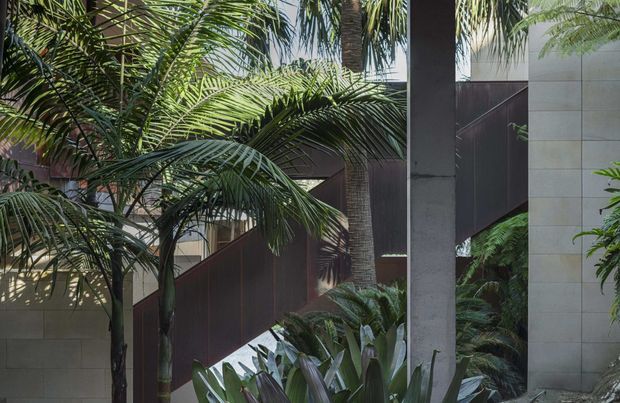 A lush tropical courtyard landscape framed by a contemporary building. Dense palm and broad-leaf planting surrounds a dark metal privacy screen, pale tiled or stone-clad walls, and a prominent raw concrete structural column. The composition has a secluded, resort-like atmosphere with layered greenery and strong vertical architectural elements.