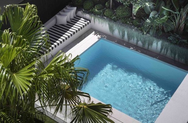 A compact courtyard-style swimming pool surrounded by lush tropical planting. The pool has pale blue water and light paving, with a raised garden bed along the rear wall. A striped black-and-white outdoor daybed or lounge seating area sits beside the pool, creating a contemporary resort-like outdoor retreat.