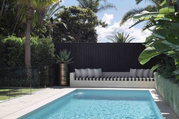 A contemporary outdoor swimming pool surrounded by lush tropical landscaping, with a built-in white lounge bench against a tall dark vertical-panel privacy fence. Frameless glass pool fencing, pale paving, sculptural planter, palms, and broad-leaf greenery create a refined resort-like setting.