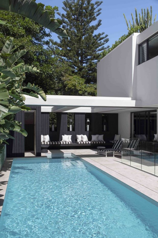A bright contemporary courtyard featuring a long rectangular swimming pool, white rendered architectural walls, dark vertical timber-look cladding, and extensive glass glazing. The pool terrace includes built-in cushioned seating and striped sun loungers, with lush tropical and mature evergreen planting surrounding the space.