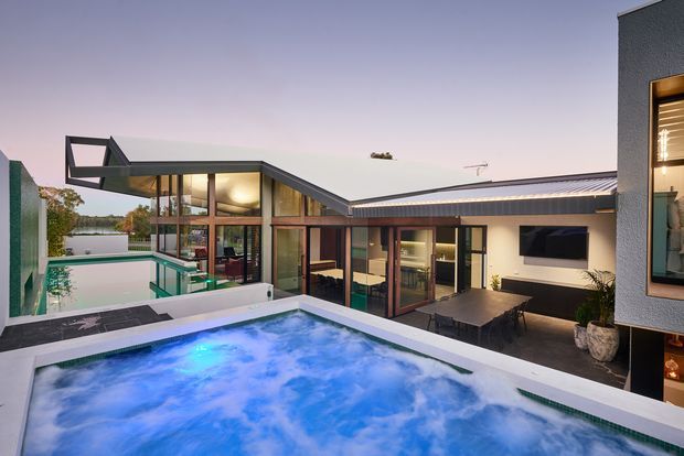 Twilight exterior view of a contemporary single-storey residence arranged around multiple swimming pools and a terrace. The home features strong geometric volumes, extensive floor-to-ceiling glazing, dark metal-framed windows, a low-pitched roof, and a sheltered outdoor entertaining area with dining furniture. A rooftop or raised foreground pool dominates the image, with a second pool visible beside the glazed living spaces.