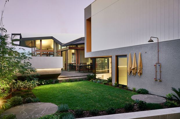 Exterior view of a contemporary multi-wing residence enclosing a landscaped courtyard. The home features crisp geometric volumes, a low-pitched roof, expansive glazing with dark timber or metal framing, light vertical panel cladding, textured grey rendered walls, and a small paved garden path with outdoor shower fixtures and hanging towels.