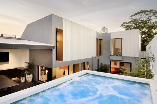 Modern multi-level residence viewed from an elevated outdoor terrace, with crisp white and grey rectilinear volumes, vertical ribbed cladding, expansive glazing, warm-lit interiors, and a prominent rooftop swimming pool in the foreground. Mature trees and planting surround the property.