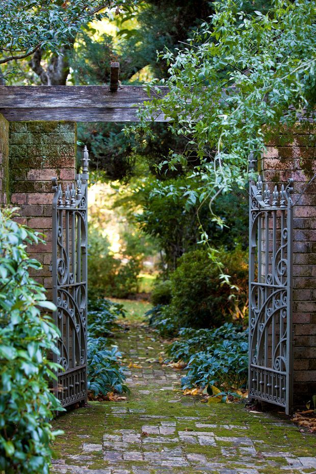 A secluded garden entrance framed by weathered brick pillars and a timber lintel, with ornate open metal gates leading onto a moss-covered stone and brick path. Dense mature greenery surrounds the pathway, creating a leafy, historic garden setting.