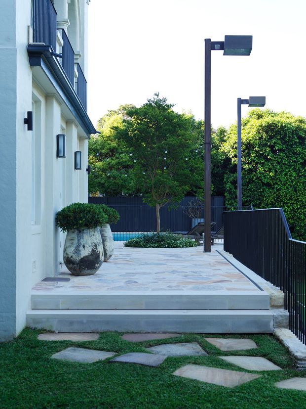 A landscaped exterior terrace beside a pale rendered building, with broad stone-paved steps and platform, dark metal balustrades and contemporary rectangular garden lights. Sculpted greenery, a large planter, stepping stones through lawn, and a glimpse of a swimming pool create a refined modern outdoor setting.