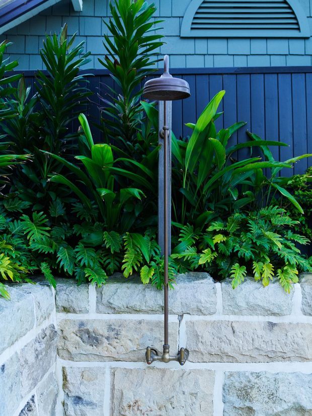 Outdoor shower with a brushed metal pipe and round showerhead mounted against a blue weatherboard-clad wall. Dense tropical planting surrounds a pale rough-cut stone retaining wall, with a louvred vent visible high on the wall.