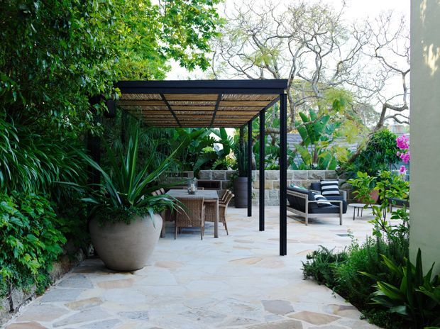 A lush landscaped outdoor courtyard or terrace with an irregular natural-stone paved floor, a slim black metal pergola with a woven or slatted roof, and outdoor dining and lounge furniture. Dense tropical and subtropical planting surrounds the space, with raised stone garden walls and a bright, leafy atmosphere.
