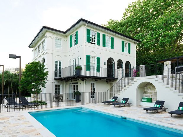 Exterior view of a grand white multi-storey residence surrounding a swimming pool. The building features classical detailing, tall arched openings, black metal railings, projecting balconies, and bright green shutters. A paved pool terrace with lounge chairs occupies the foreground, with mature trees and a distant urban backdrop.