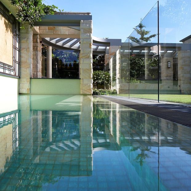 A modern residential pool courtyard viewed at water level, featuring pale natural stone columns and walls, dark horizontal roof and pergola elements, extensive glass pool fencing, and lush greenery. The clear blue pool dominates the foreground and reflects the surrounding architecture and sky.